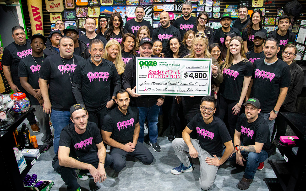 The entire Ooze Wholesale team stands in black t shirts with the pink Ooze logo in the Ooze showroom. They surround a giant donation check made out to the Shades of Pink Foundation, held in the center by Foundation Executive Director Karla Sherry