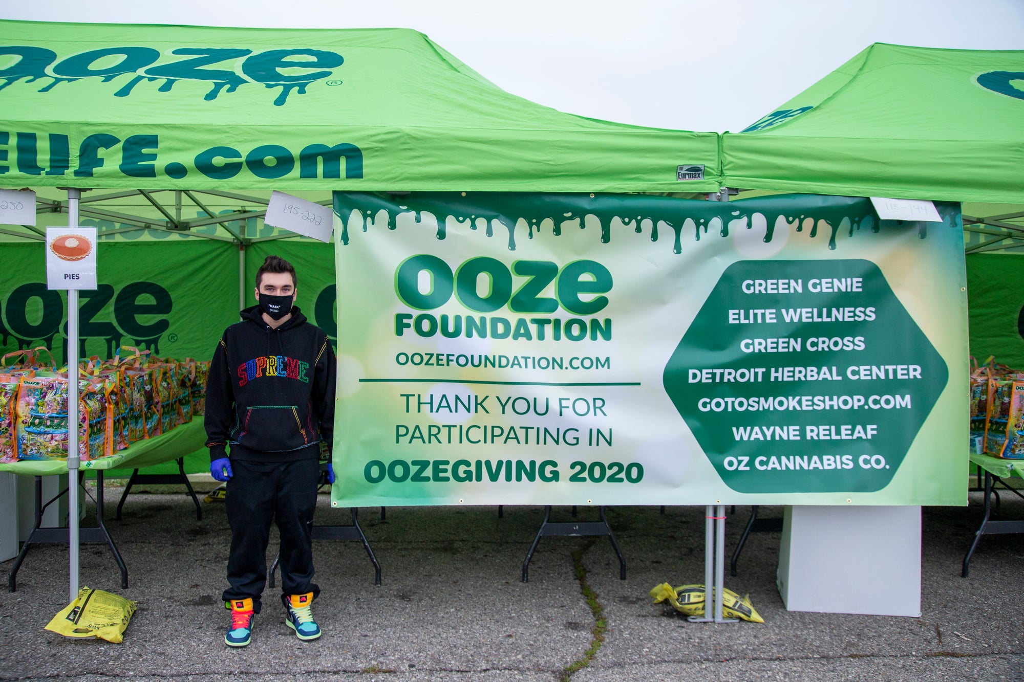 A young man wearing a black face mask and black track suit stands next to the Ooze Foundation Oozegiving banner. He is under the green Ooze tent in front of the donation gift bags.
