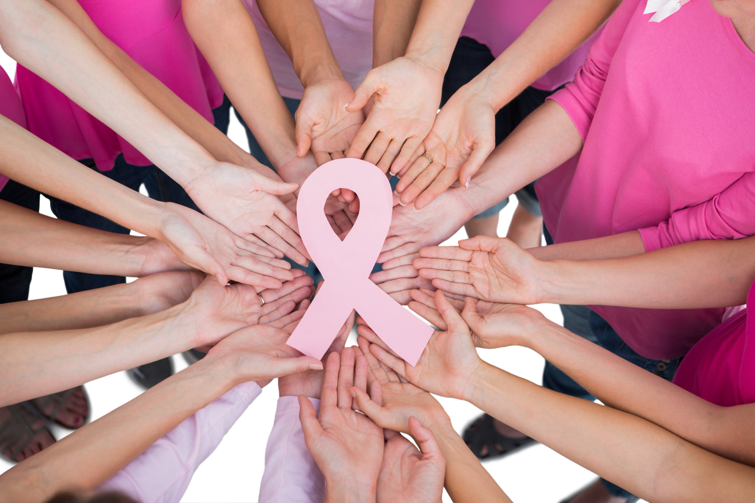 A group of women of different ages and races are gathered together in a circle. They all have their arms stretched out to hold up the light pink breast cancer ribbon in the center. They are all wearing different shades of pink shirts.
